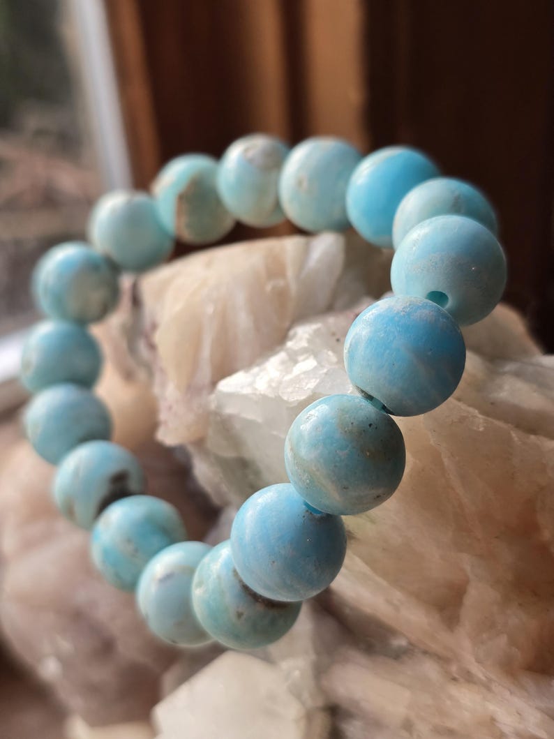 May include: A beaded bracelet featuring round, light blue beads. The beads have a slightly mottled appearance with variations in color. The bracelet is strung together, creating a circular shape. The background is a light-colored, textured surface.