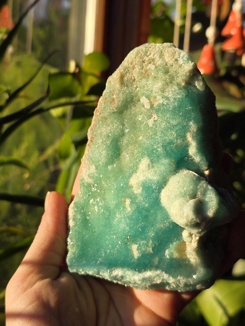 Puede incluir: Una mano sostiene un esp&eacute;cimen mineral de color turquesa con bordes &aacute;speros. La piedra tiene una superficie texturizada con parches m&aacute;s claros y una peque&ntilde;a protuberancia redondeada. El fondo est&aacute; borroso, con toques de follaje verde y luz solar.