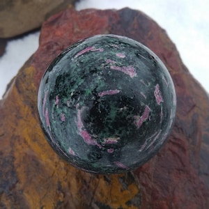 May include: A green and pink gemstone sphere with a polished surface. The sphere is resting on a brown and red rock.