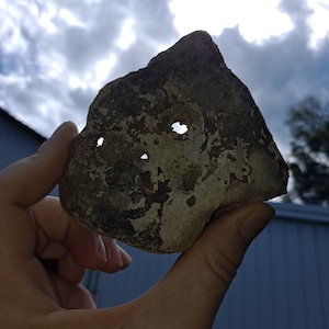 May include: A hand holds a gray rock with three holes in it. The rock is irregularly shaped and has a rough texture. The background is a cloudy sky.