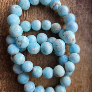 May include: Several strands of light blue gemstone beads are arranged on a wooden surface. The beads are round and have a marbled appearance, with variations in color. The beads are approximately 0.4 inches in diameter.