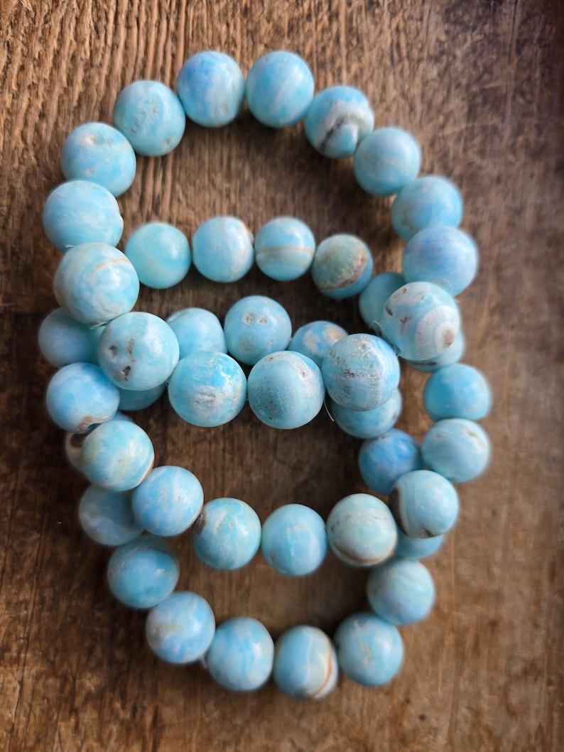 May include: Several strands of light blue gemstone beads are arranged on a wooden surface. The beads are round and have a marbled appearance, with variations in color. The beads are approximately 0.4 inches in diameter.