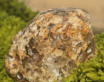 May include: A large, rough, clear quartz crystal with a brown, cloudy center. The crystal is sitting on a bed of green moss.