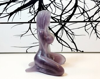 May include: A purple glass figurine of a woman kneeling with her head tilted down. The figurine is translucent and has a smooth, polished finish.