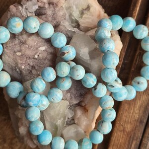 May include: Several strands of light blue gemstone bead bracelets. The beads are round and have a marbled appearance with darker blue and brown inclusions. The bracelets are resting on a light-colored, textured stone and a wooden surface.