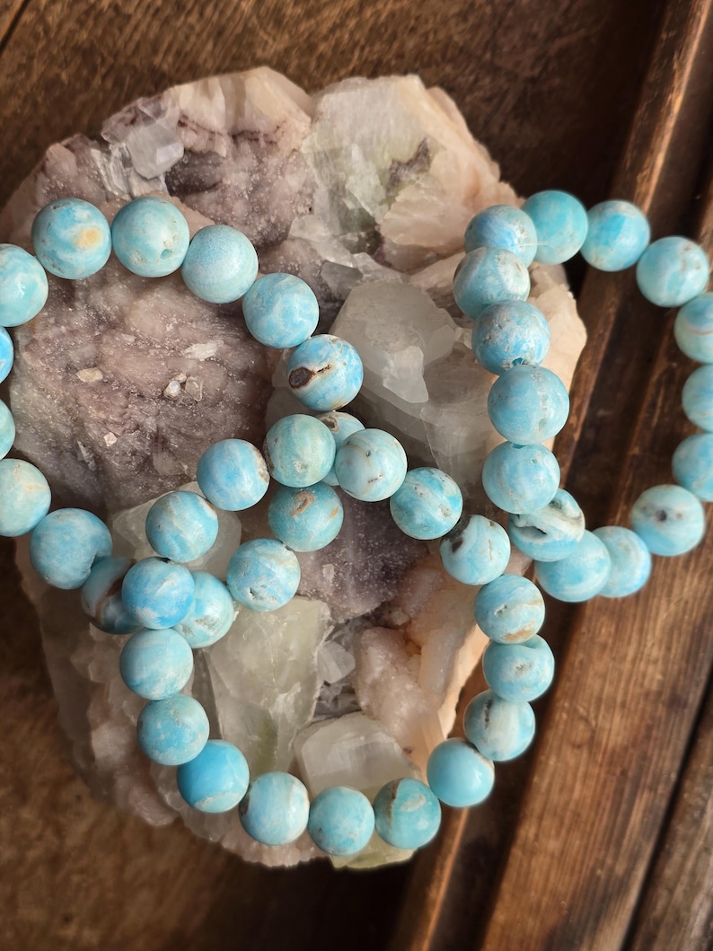 May include: Several strands of light blue gemstone bead bracelets. The beads are round and have a marbled appearance with darker blue and brown inclusions. The bracelets are resting on a light-colored, textured stone and a wooden surface.
