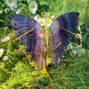 Peut inclure: Un cristal de fluorine en forme de papillon violet et or sur un support en métal doré. Le cristal est posé sur un lit de mousse verte.