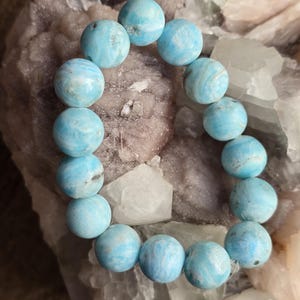 May include: A bracelet made of round, light blue beads with white marbling. The beads are strung together, forming a circular shape. The bracelet is displayed on a rough, light-colored stone surface.