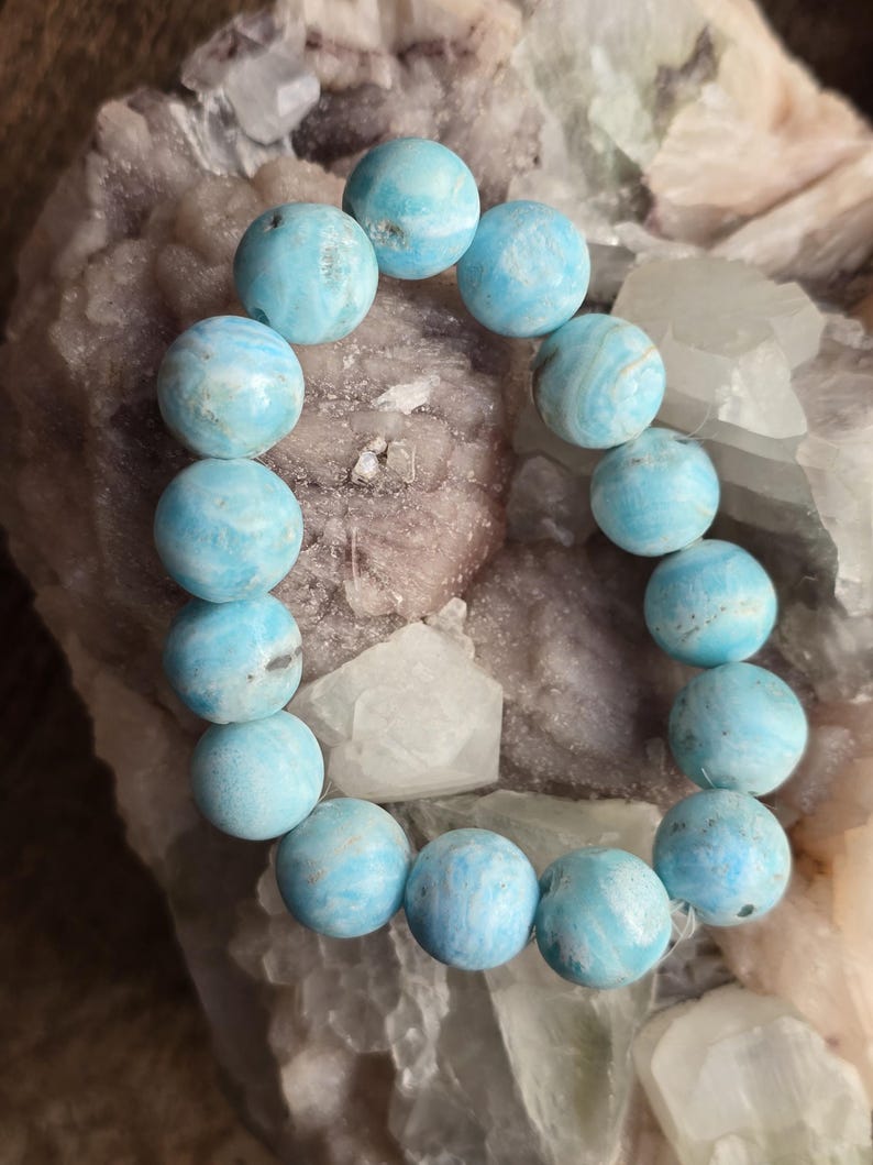 May include: A bracelet made of round, light blue beads with white marbling. The beads are strung together, forming a circular shape. The bracelet is displayed on a rough, light-colored stone surface.