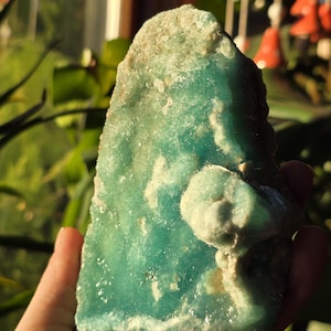 Puede incluir: Una mano sostiene un esp&eacute;cimen mineral en bruto de color turquesa. La piedra tiene una apariencia en capas con una superficie m&aacute;s clara y texturizada en la parte superior. Una formaci&oacute;n redondeada y de color m&aacute;s claro sobresale del lado derecho.