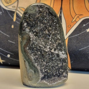 May include: A green and black crystal geode with a rough exterior and a sparkly, crystal-lined interior. The geode is shaped like a teardrop and is about 12.5 cm tall.