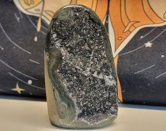 May include: A green and black crystal geode with a rough exterior and a sparkly, crystal-lined interior. The geode is shaped like a teardrop and is about 5 inches tall.