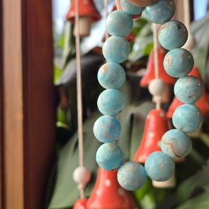 May include: A bracelet of round, light blue beads with subtle marbling. The beads are held in a hand, with red mushroom-shaped decorations in the background. The bracelet is a light blue color.