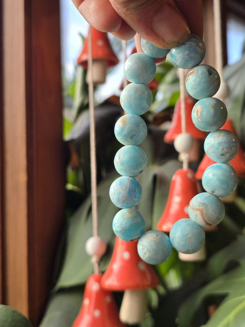 May include: A bracelet of round, light blue beads with subtle marbling. The beads are held in a hand, with red mushroom-shaped decorations in the background. The bracelet is a light blue color.
