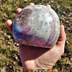 May include: A large, smooth, polished, purple and white gemstone with swirling patterns. The stone is held in a hand.