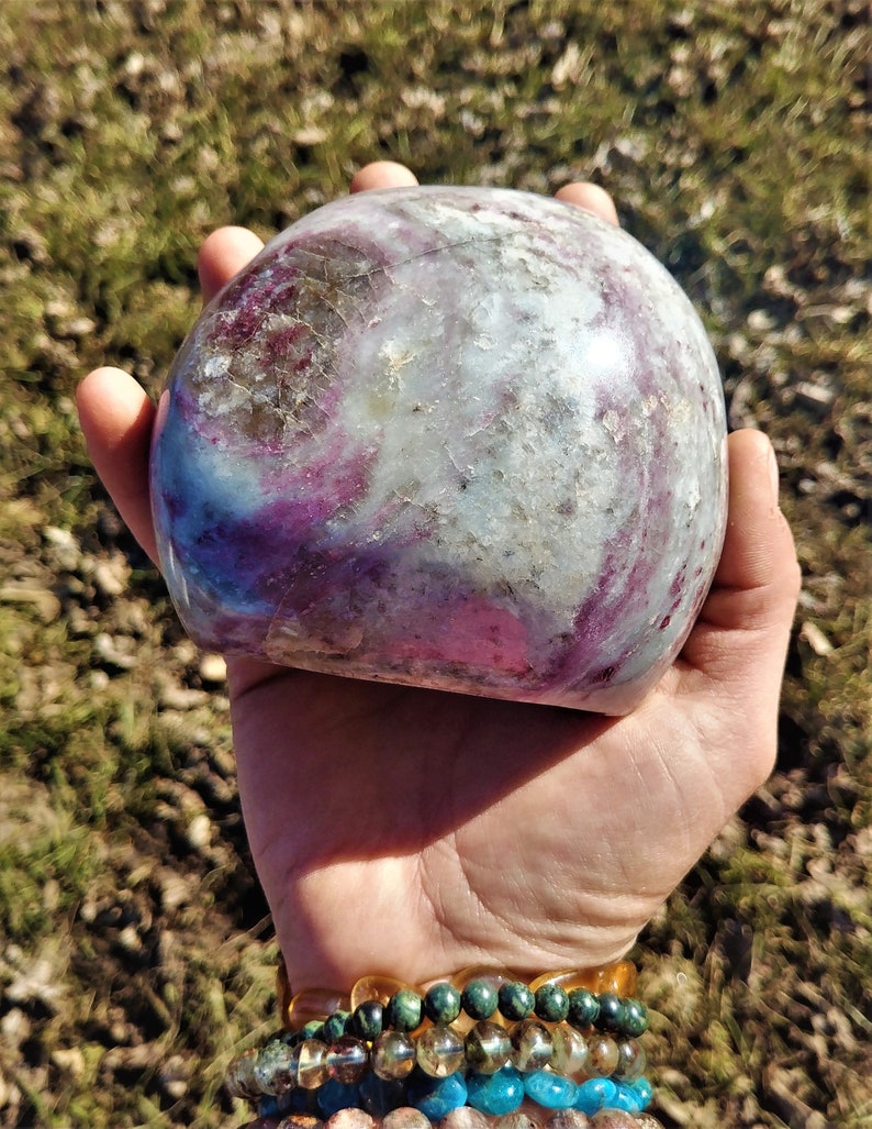 May include: A large, smooth, polished, purple and white gemstone with swirling patterns. The stone is held in a hand.