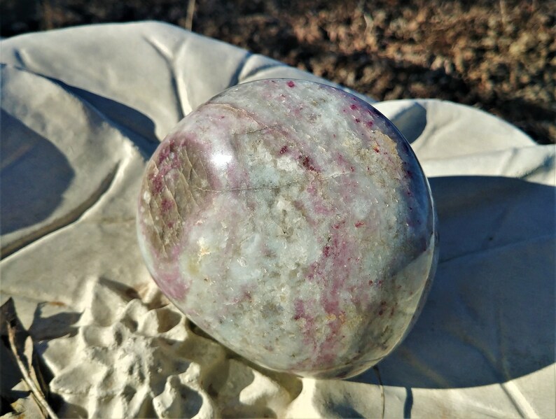 May include: A polished, round, white and pink gemstone with a smooth, shiny surface. The gemstone has a swirling pattern of pink and white.
