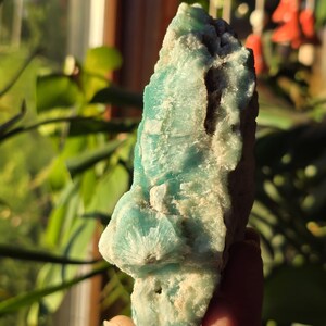 Puede incluir: Una gema natural en bruto con una llamativa paleta de colores turquesa y blanco. La piedra tiene una superficie rugosa y texturizada con formaciones cristalinas. La forma general es irregular, mostrando la belleza natural del mineral. La gema se sostiene en una mano.