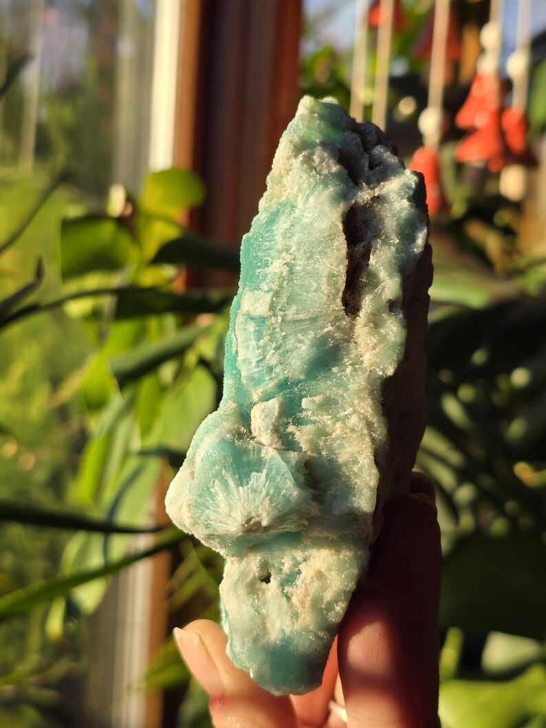 Puede incluir: Una gema natural en bruto con una llamativa paleta de colores turquesa y blanco. La piedra tiene una superficie rugosa y texturizada con formaciones cristalinas. La forma general es irregular, mostrando la belleza natural del mineral. La gema se sostiene en una mano.