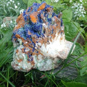 May include: A large, rough, natural crystal cluster with blue, green, white, and orange hues. The crystal is surrounded by green grass and white flowers.
