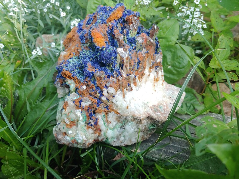 May include: A large, rough, natural crystal cluster with blue, green, white, and orange hues. The crystal is surrounded by green grass and white flowers.