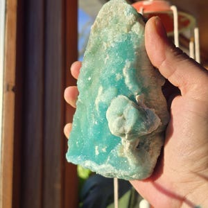 Puede incluir: Un esp&eacute;cimen mineral en bruto de color turquesa con una estructura cristalina. La piedra tiene un tono azul claro con &aacute;reas blancas y una textura ligeramente rugosa. El mineral se sostiene en una mano, con una ventana y elementos decorativos en el fondo.