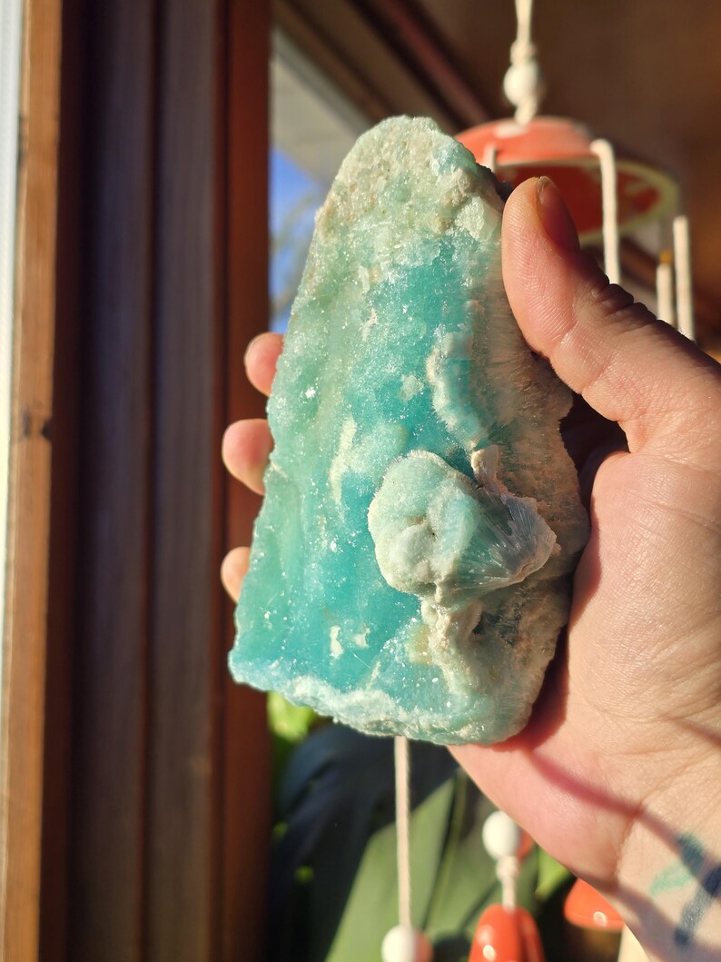 Puede incluir: Un esp&eacute;cimen mineral en bruto de color turquesa con una estructura cristalina. La piedra tiene un tono azul claro con &aacute;reas blancas y una textura ligeramente rugosa. El mineral se sostiene en una mano, con una ventana y elementos decorativos en el fondo.