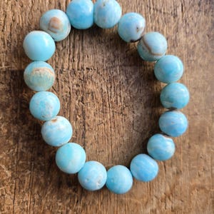 May include: A bracelet made of round, light blue beads with subtle variations in color. The beads are strung together, forming a circular shape. The bracelet is displayed on a wooden surface, providing a natural backdrop.