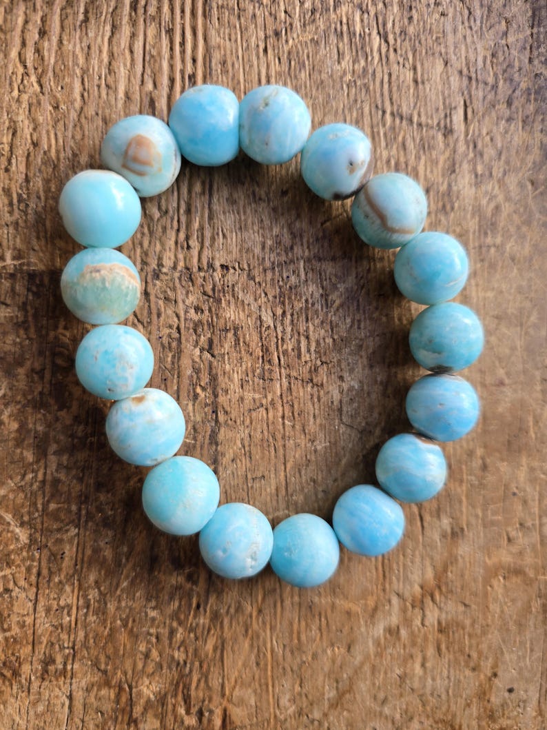 May include: A bracelet made of round, light blue beads with subtle variations in color. The beads are strung together, forming a circular shape. The bracelet is displayed on a wooden surface, providing a natural backdrop.