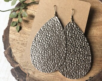 May include: A pair of silver teardrop earrings with a textured, hammered metal appearance. The earrings hang from gold-coloured hooks and are displayed on a brown craft paper card, resting on a wooden slice.