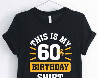 Peut inclure: T-shirt noir avec l'inscription "THIS IS MY 60th BIRTHDAY SHIRT" en blanc et jaune. Le t-shirt est suspendu à un cintre sur fond blanc. Le texte est centré sur le devant du t-shirt.