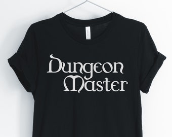 May include: Black t-shirt with the words "Dungeon Master" in white, stylized font. The shirt is hanging on a metal hanger against a white background. The sleeves are cuffed.