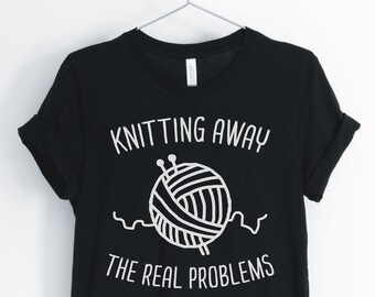 Puede incluir: Camiseta negra con texto blanco que dice "Knitting Away The Real Problems" y un gráfico de un ovillo de lana con agujas de tejer.