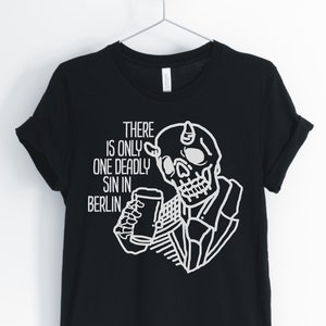 Puede incluir: Camiseta negra con un gr&aacute;fico blanco de un esqueleto con cuernos bebiendo de un vaso. El texto "THERE IS ONLY ONE DEADLY SIN IN BERLIN" est&aacute; impreso en la camiseta.