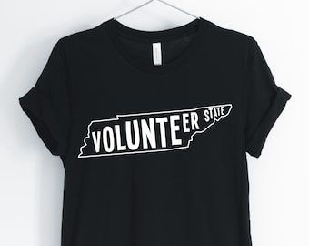 Puede incluir: Camiseta negra con un contorno blanco del estado de Tennessee y el texto "Volunteer State" dentro del contorno.