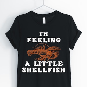 May include: Black t-shirt with white text that reads "I'm Feeling A Little Shellfish" and a drawing of a lobster in orange.