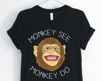 Op de afbeelding: Zwart T-shirt met een witte afbeelding van een aap met de tekst "Monkey See Monkey Do"
