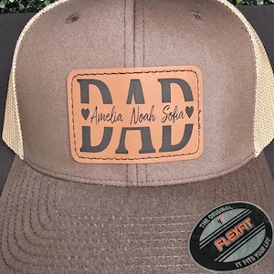 May include: A brown trucker hat with a tan mesh back. The front features a leather patch with the word "DAD" in bold letters, and the names "Amelia Noah Sofia" are below. The hat has a curved brim and a Flexfit tag.