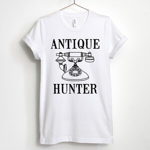 May include: White t-shirt with black text that reads "Antique Hunter" and a black and white illustration of an antique rotary phone.
