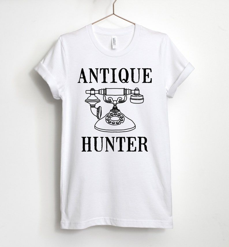 May include: White t-shirt with black text that reads "Antique Hunter" and a black and white illustration of an antique rotary phone.