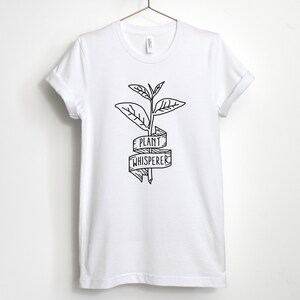 Puede incluir: Camiseta blanca con un dibujo en l&iacute;nea negra de una planta y una pancarta que dice "Plant Whisperer".