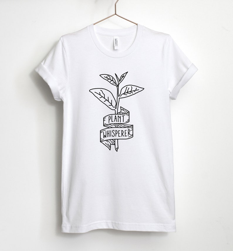 Puede incluir: Camiseta blanca con un dibujo en l&iacute;nea negra de una planta y una pancarta que dice "Plant Whisperer".
