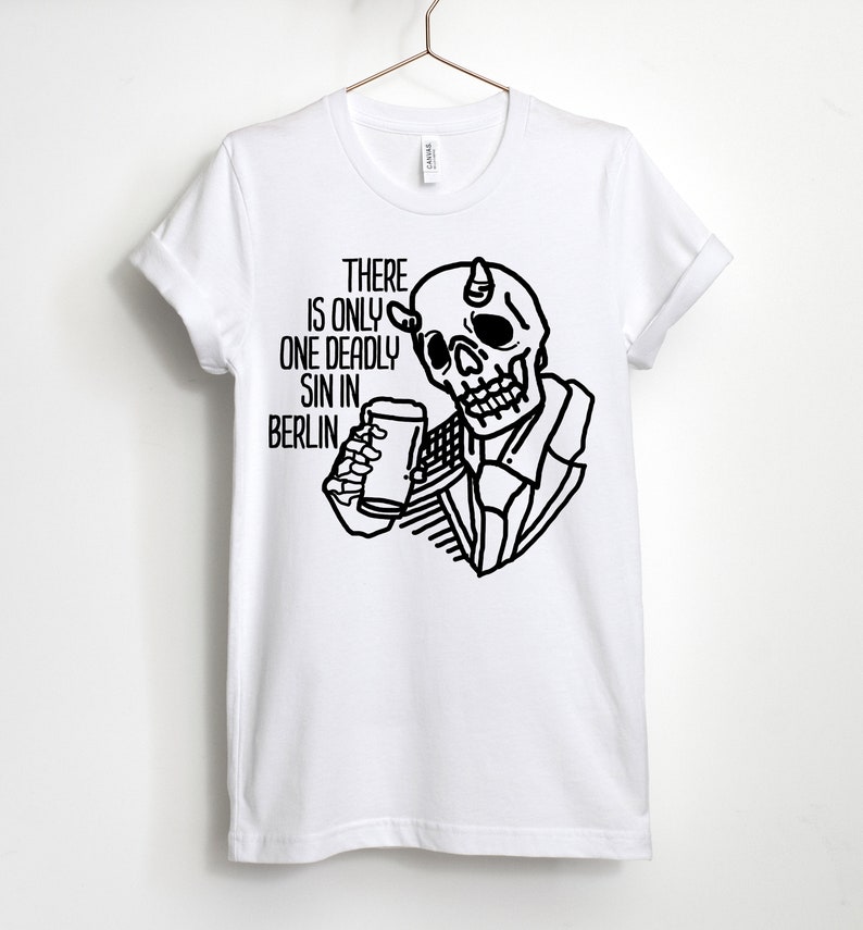 Puede incluir: Camiseta blanca con una ilustraci&oacute;n en blanco y negro de una calavera con cuernos sosteniendo un vaso de cerveza. El texto "THERE IS ONLY ONE DEADLY SIN IN BERLIN." est&aacute; impreso en la camiseta.