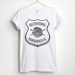 May include: White t-shirt with a black graphic of a yarn ball and knitting needles inside a shield with the text "RECOVERING YARNAHOLIC".