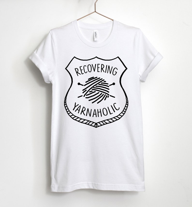 May include: White t-shirt with a black graphic of a yarn ball and knitting needles inside a shield with the text "RECOVERING YARNAHOLIC".
