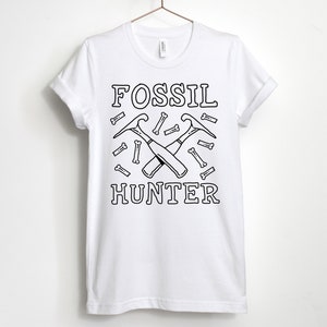 May include: White t-shirt featuring a black graphic design of two crossed pickaxes and the words "FOSSIL HUNTER" surrounded by small bone shapes.