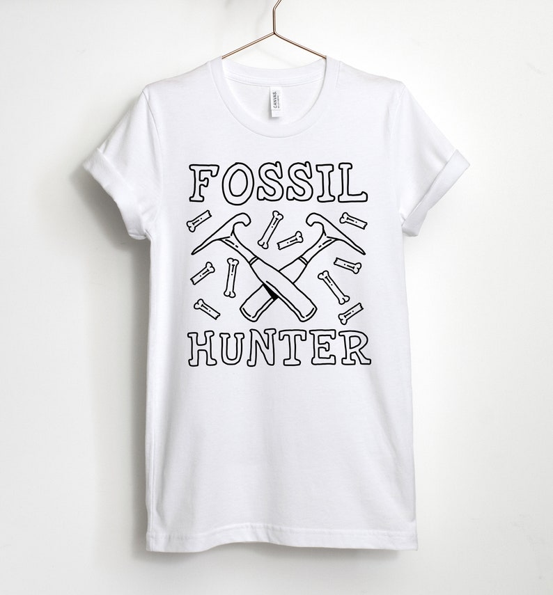 May include: White t-shirt featuring a black graphic design of two crossed pickaxes and the words "FOSSIL HUNTER" surrounded by small bone shapes.