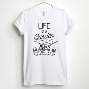 May include: White t-shirt with black text that reads "Life is a Garden, Dig It."  The text is accompanied by a black and white illustration of a wheelbarrow with gardening tools.