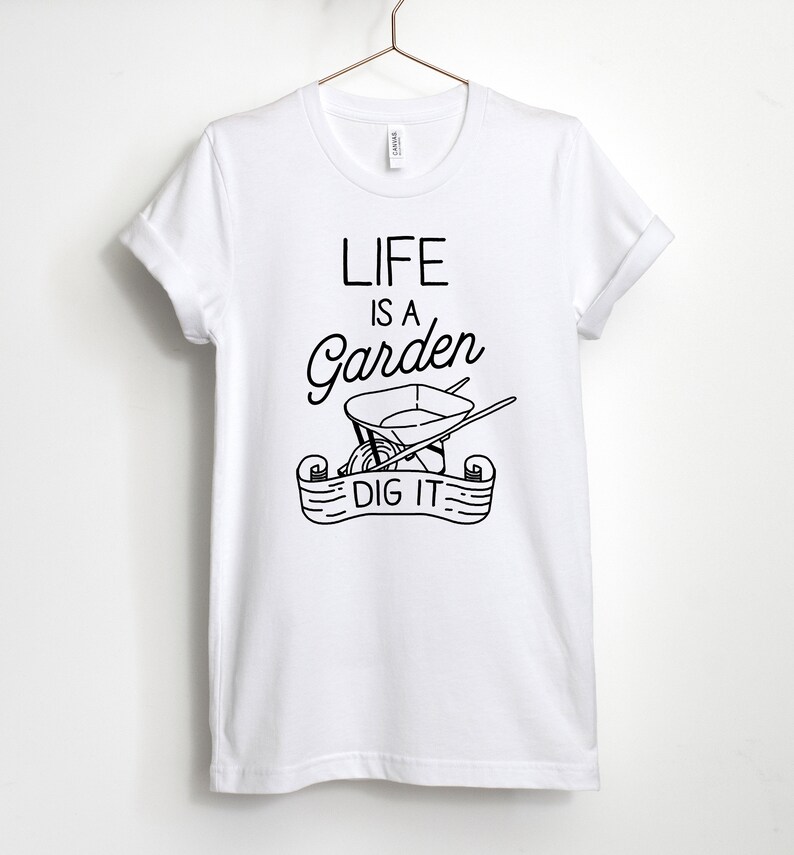 May include: White t-shirt with black text that reads "Life is a Garden, Dig It."  The text is accompanied by a black and white illustration of a wheelbarrow with gardening tools.