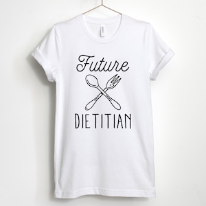 K&ouml;nnte beinhalten: Wei&szlig;es T-Shirt mit schwarzem Text "Future Dietitian" und einer Grafik einer gekreuzten Gabel und L&ouml;ffel.